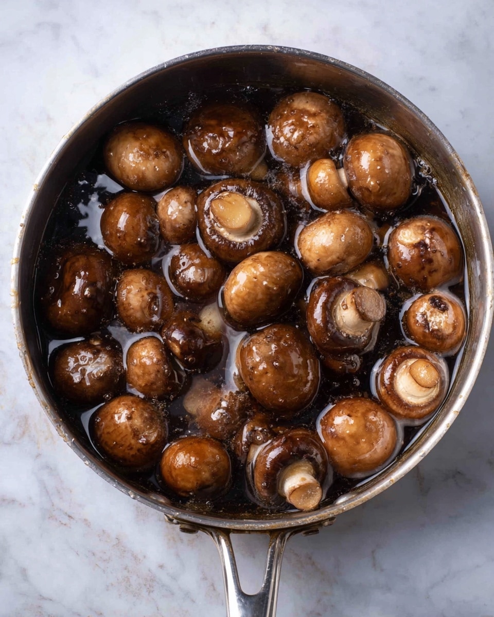 A silver metal pan filled with whole brown mushrooms submerged in dark water with small bubbles on the surface; the mushrooms have smooth, shiny caps varying in size and some show the lighter underside with gills; the pan rests on a white marbled surface, and the handle of the pan is visible at the bottom center of the image photo taken with an iphone --ar 4:5 --v 7