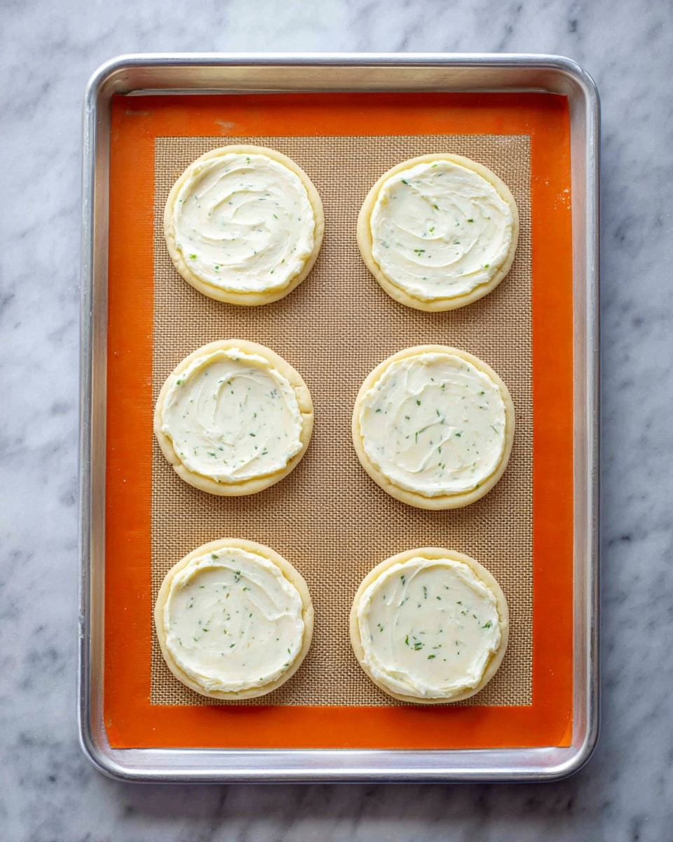 The image shows six small round dough bases arranged in two vertical columns on a silver baking tray lined with an orange silicone baking mat. Each dough base is topped with a smooth, creamy white layer of spread with subtle green herb flecks, evenly covering the center inside a slightly raised dough crust. The surface beneath the tray is a white marbled texture. The scene is viewed from above in clear, bright lighting. photo taken with an iphone --ar 4:5 --v 7
