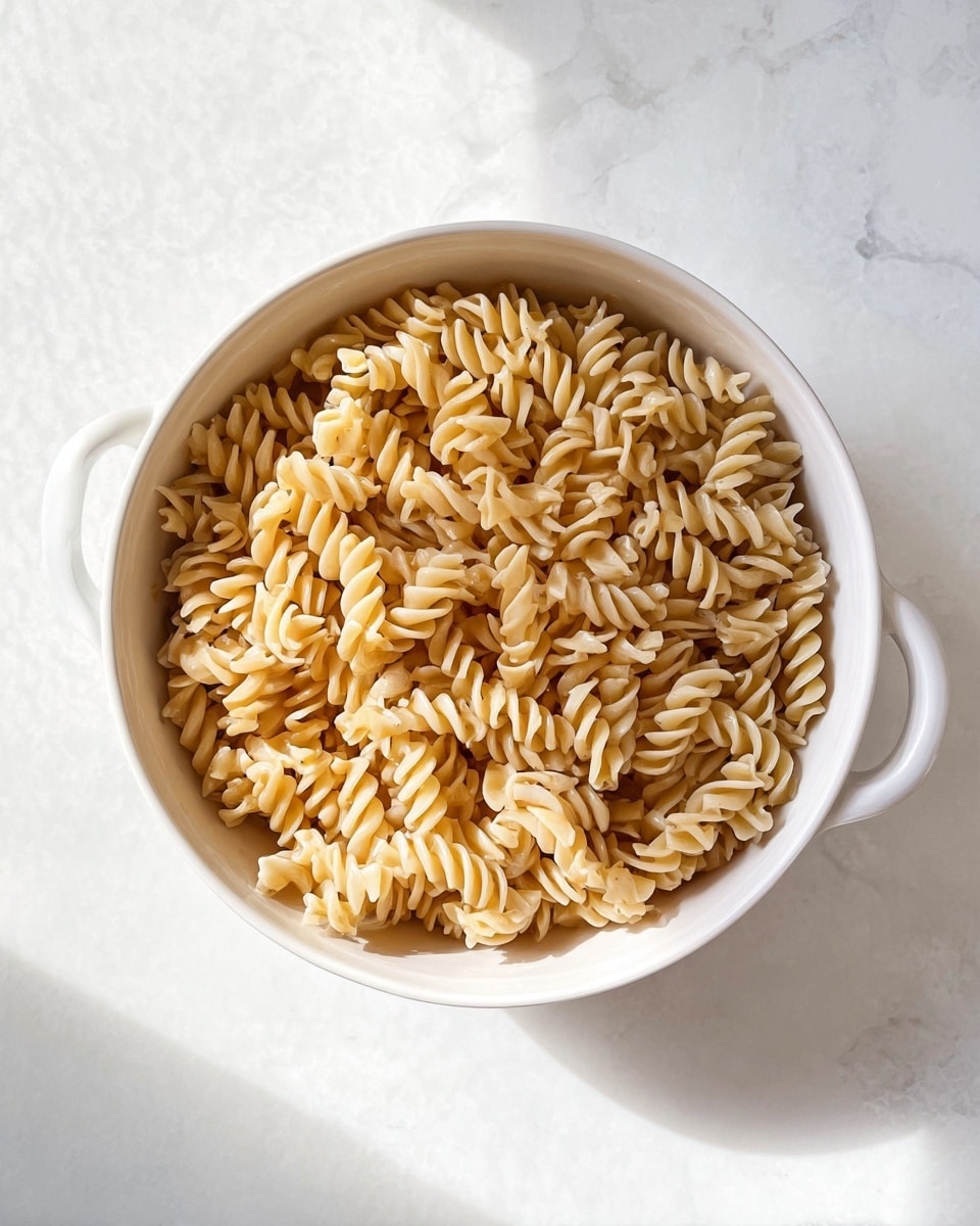 A white bowl with two handles is filled with a single layer of cooked spiraled pasta, showing a light beige color with a slightly glossy texture. The bowl sits on a white marbled surface with soft shadows around it, and the image is taken from directly above, capturing the pasta pieces packed closely but clearly visible individually photo taken with an iphone --ar 4:5 --v 7