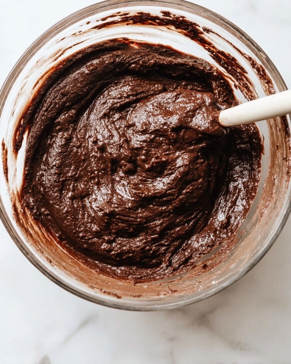 Healthy Chocolate Avocado Muffins for Kids Recipe 5 A clear glass bowl holds thick dark brown chocolate batter with a slightly rough texture. A mixing spatula is partially visible, blending the thick batter inside the bowl. The bowl sits on a white marbled surface, with some batter spread on the inside edges of the bowl. The mixture looks dense and rich, with small lumps giving a textured feel. Photo taken with an iphone --ar 4:5 --v 7