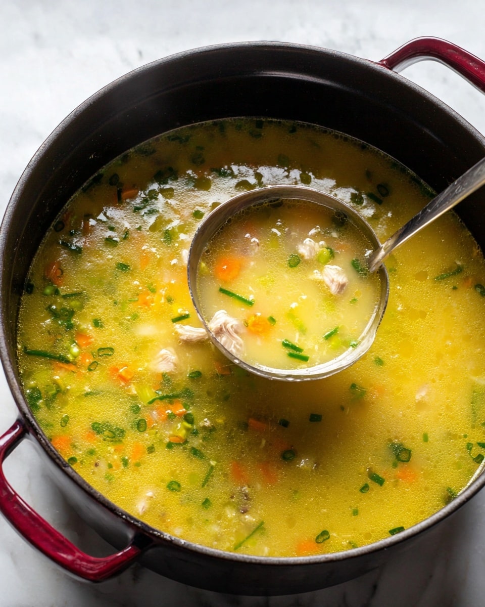 A bowl with a thin blue rim and dark edge holds a clear chicken soup filled with shredded chicken pieces, small chopped bright orange carrots, light green celery bits, and finely chopped green onions. The soup has a yellow broth with good clarity and scattered fresh green cilantro leaves on top. There is a slice of lemon placed on one side inside the soup, with some black pepper sprinkled across the surface. In the background, there is a smaller white bowl with lemon wedges on a white marbled surface and a fringed white linen cloth partially underneath the bowl. Photo taken with an iphone --ar 4:5 --v 7