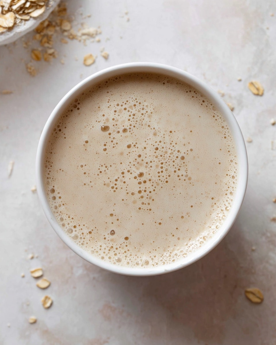 The image shows the inside of a blender from above with several ingredients layered. At the bottom, there is a whole raw egg yolk surrounded by thick white liquid likely milk or yogurt. Around the edges, there are scattered light brown rolled oats. Some areas show a swirl of dark brown liquid, possibly syrup or vanilla extract mixing with the white liquid. The blender container is transparent, placed on a white marbled surface with a few rolled oats scattered around. Photo taken with an iphone --ar 4:5 --v 7