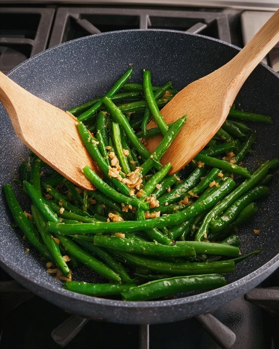 A white scalloped plate holds bright green cooked green beans arranged in a small pile. A topping of finely chopped, golden-brown crispy garlic is spread evenly across the top center of the green beans, adding texture and color contrast. The plate sits on a white marbled surface, and a light pink cloth napkin is placed to the lower left side. To the right of the plate, pink-handled chopsticks rest on the surface next to a translucent glass with a pinkish drink inside. The scene is softly lit, highlighting the fresh green and golden tones. photo taken with an iphone --ar 4:5 --v 7