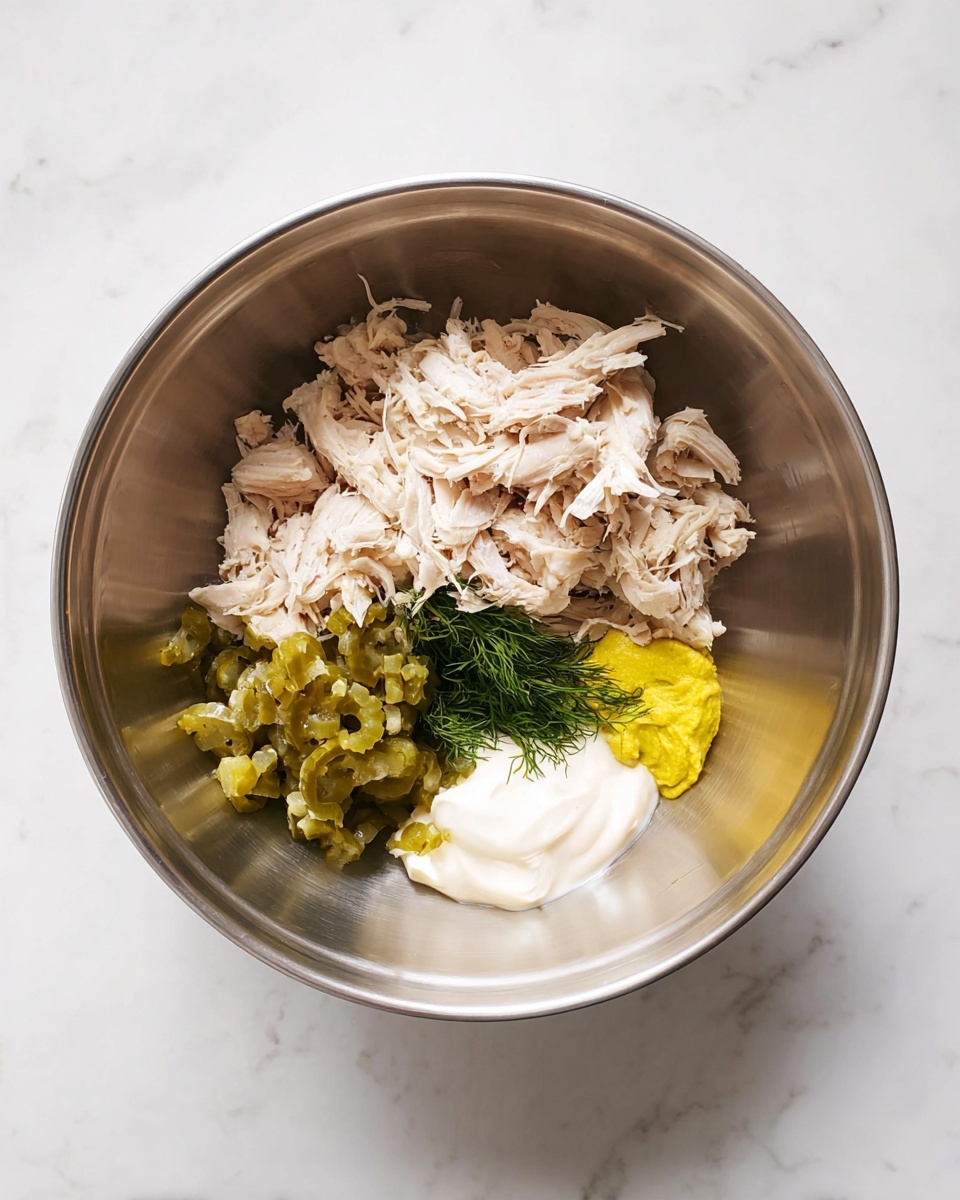 Inside a shiny metal bowl, shredded light beige chicken pieces take up most of the space, with small piles of chopped green pickles on one side and a dollop of thick white mayonnaise below them. Next to the mayonnaise, there is a small bright yellow patch of mustard, and a tiny bunch of fresh green dill sits on the edge near the pickles. The bowl is placed on a white marbled surface with soft veins running through it. photo taken with an iphone --ar 4:5 --v 7